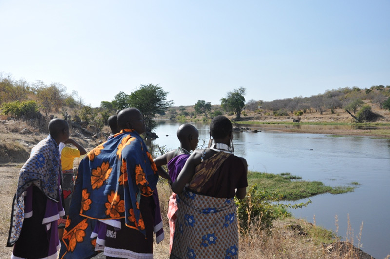 A group of people in colorful patterned clothing stand by a riverbank, looking out over the water under a clear sky, with trees and dry grassland in the background.