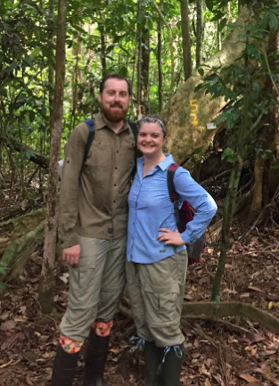 A man and woman wearing outdoor clothing and rubber boots stand together in a lush forest, smiling at the camera. The background includes trees, leaves on the ground, and a large rock marked with yellow numbers.