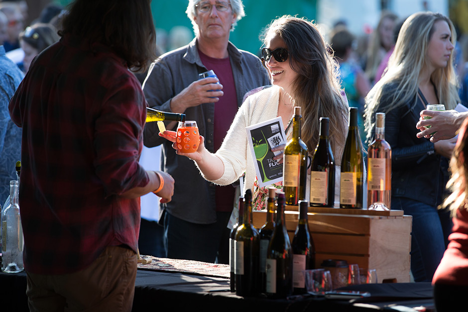 A woman wearing sunglasses smiles as she receives a wine pour from someone at an outdoor event. Several wine bottles are displayed on the table between them, and people socialize in the background.