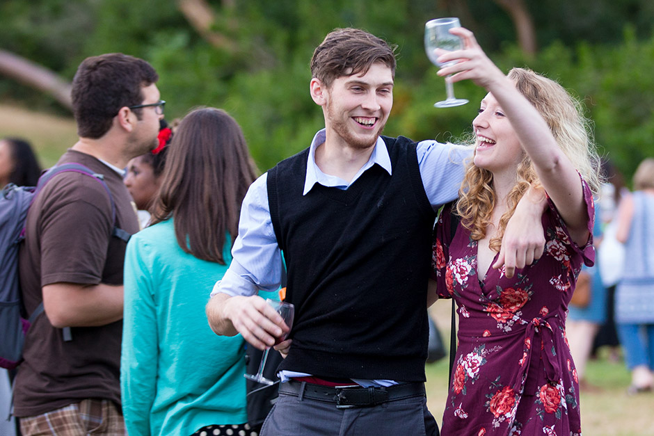 A man and a woman smile and laugh together at an outdoor gathering. The woman holds up a wine glass, and the man has his arm around her shoulders. Other people are socializing in the background.