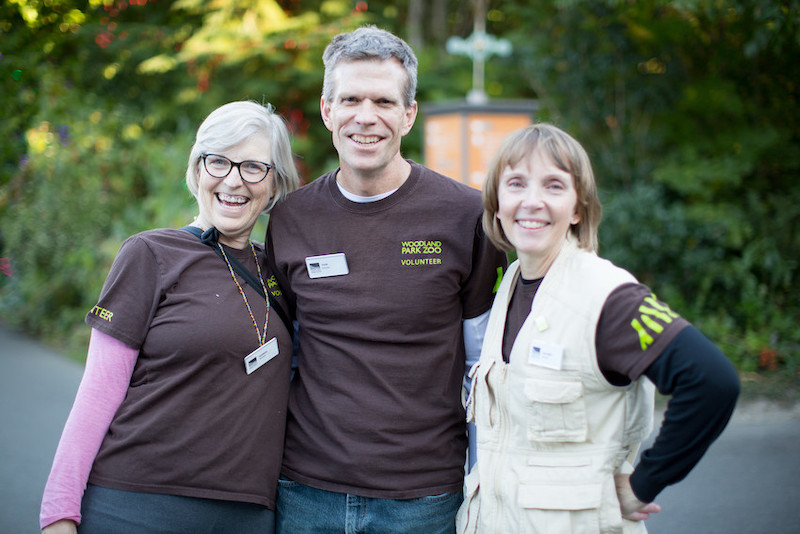 Three smiling adults, two women and one man, stand close together outdoors. They are wearing brown shirts with “Woodland Park Zoo Volunteer” logos and name badges, with trees and greenery in the background.