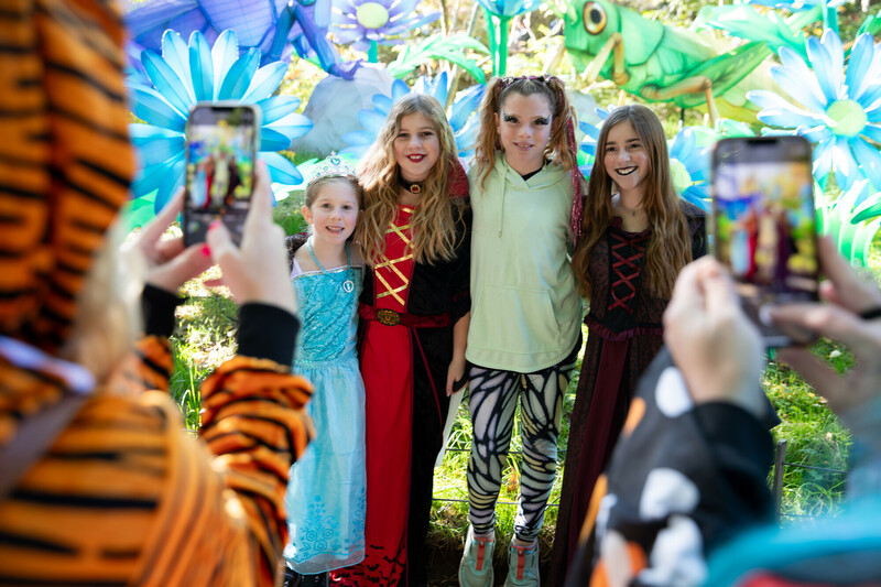 Four children in colorful costumes pose and smile for photos outdoors in front of oversized cartoon flowers and bugs. Two people in animal costumes are taking pictures with their phones in the foreground.