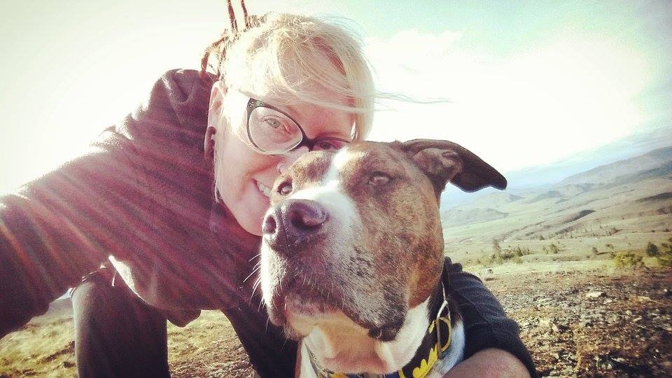 A person with glasses and blonde hair poses closely with a brown and white dog outdoors on a sunny day, with hills and open landscape in the background. Sunlight rays partially illuminate the scene.