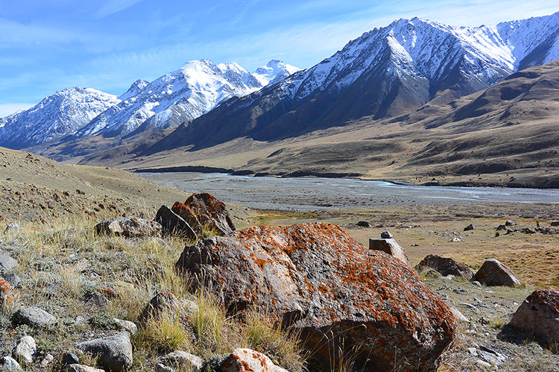 Snow-capped mountains rise in the background behind a river valley, with grassy terrain and large, orange-lichen-covered rocks in the foreground under a blue sky.
