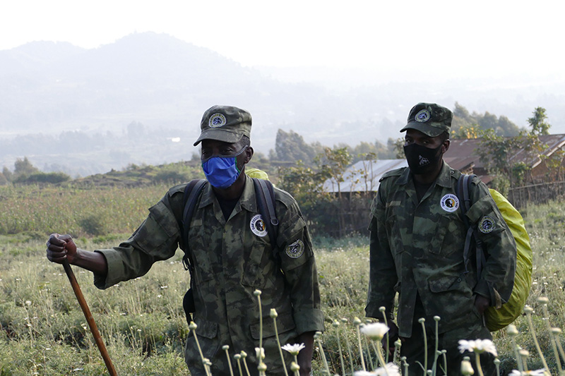 Two people in green camouflage uniforms and face masks stand in a grassy field with mountains in the background. Both wear caps and backpacks, and one holds a walking stick.