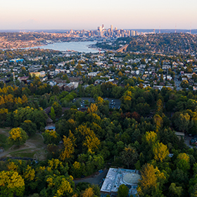Aerial view of the zoo with a dense area of trees and greenery in the foreground, urban neighborhoods in the midground, and a skyline with tall buildings in the background under a clear sky.