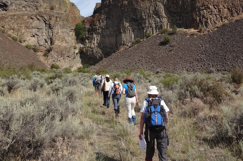 A group of people with backpacks and hats hike along a grassy trail surrounded by sagebrush, rocky hills, and cliffs under a partly sunny sky.
