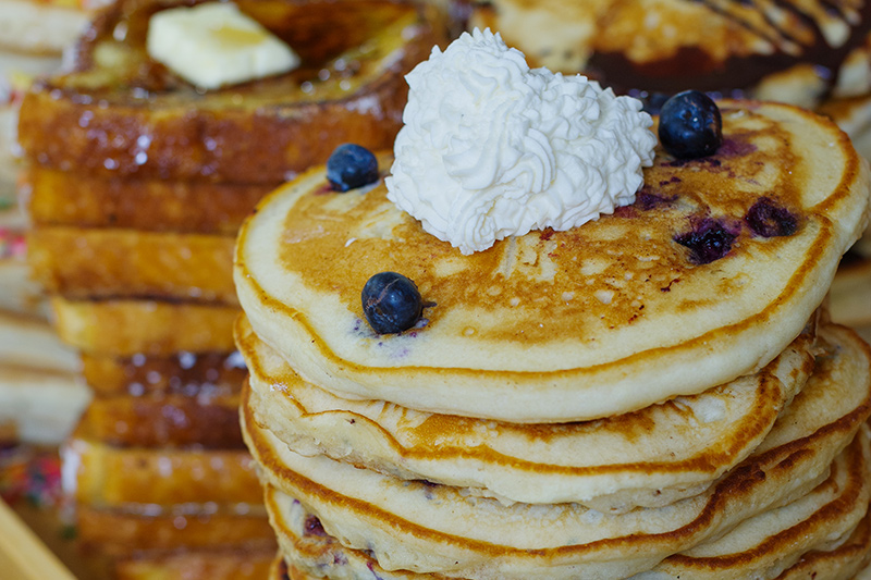 A stack of blueberry pancakes topped with whipped cream and blueberries, with slices of buttered French toast in the background.