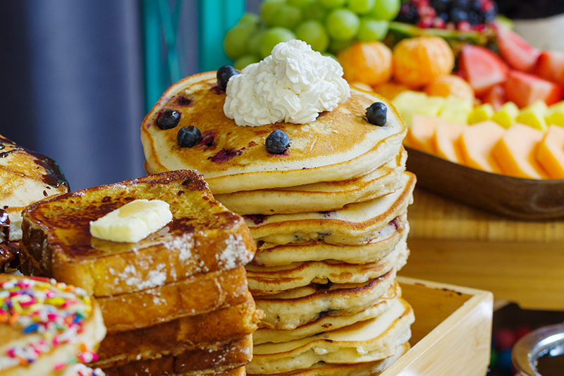 A stack of blueberry pancakes topped with whipped cream is next to slices of French toast with butter. In the background, there is an assortment of fresh fruit including grapes, melon, and berries.