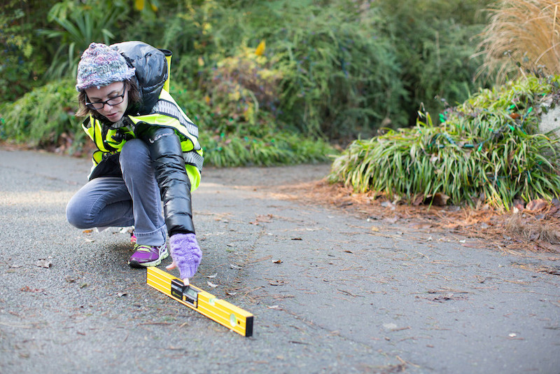 A person wearing a winter hat, gloves, and reflective vest crouches on a paved path, using a yellow spirit level to check the ground’s evenness near green plants and grass.