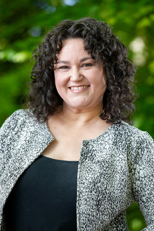 A woman with curly dark hair smiles at the camera. She is wearing a black top and a light-colored textured jacket, standing outdoors with a blurred green leafy background.