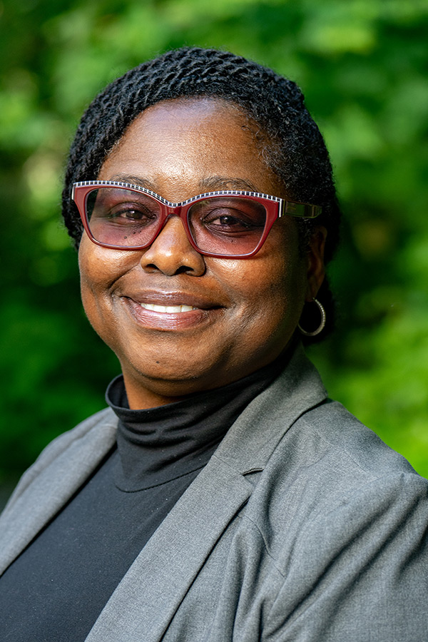 A woman with short braided hair, wearing red glasses, a gray blazer, and a black turtleneck, smiles at the camera with a green, blurred outdoor background.