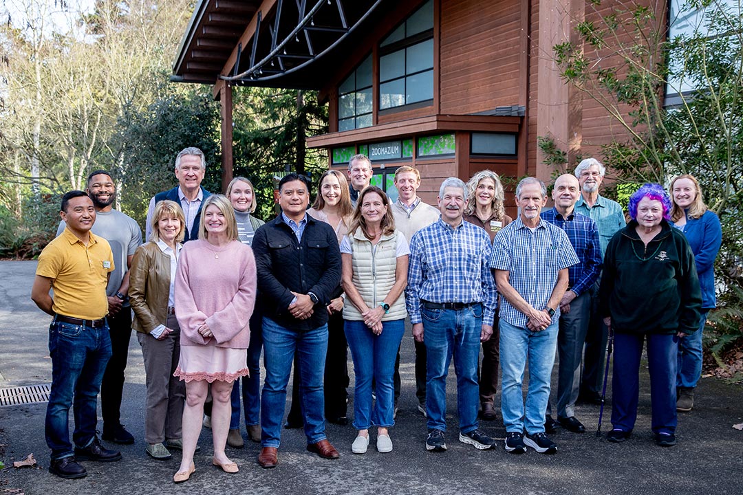 A group of seventeen adults stands outdoors in front of a wooden building, smiling at the camera. Trees and greenery surround them, and the weather appears sunny and pleasant.