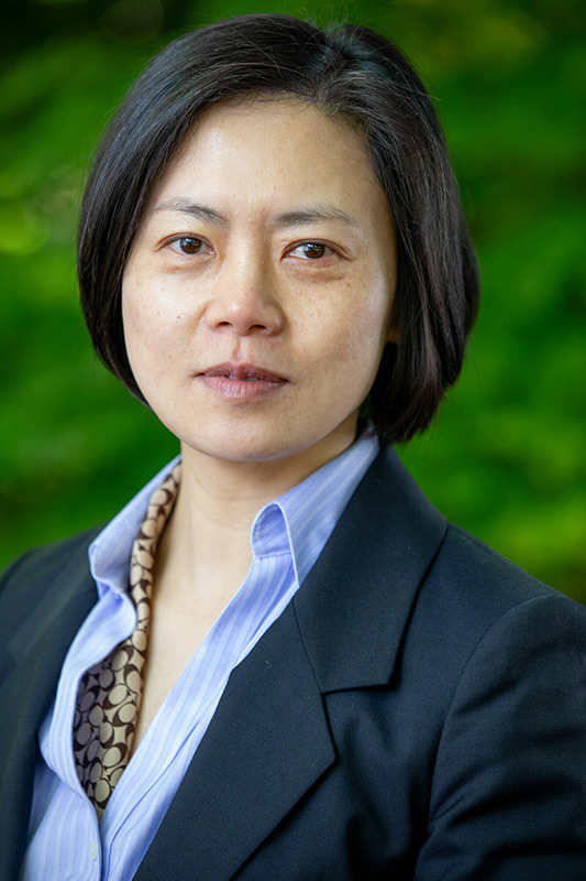 A person with short dark hair wearing a black blazer, light blue shirt, and patterned scarf stands outdoors in front of a blurred green background, looking directly at the camera with a neutral expression.