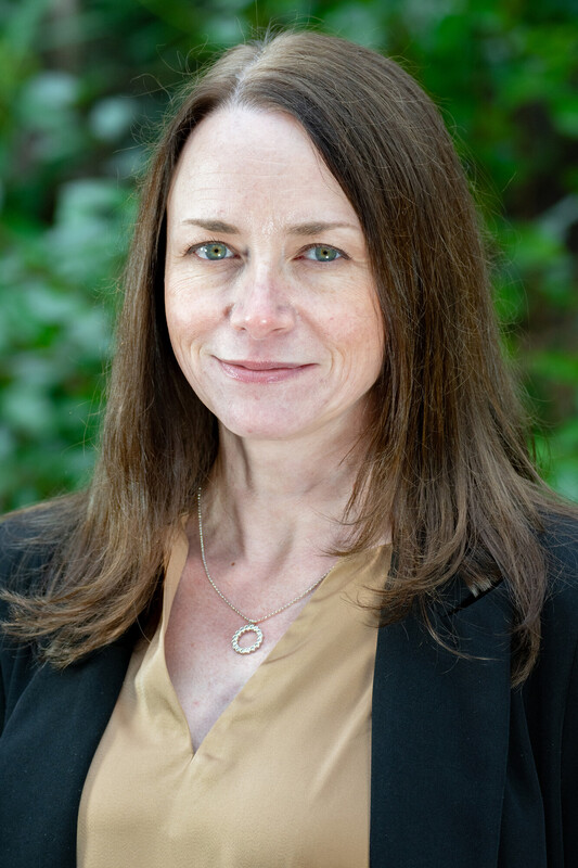 A woman with straight brown hair, wearing a black blazer over a tan blouse and a necklace, stands outdoors in front of green foliage, smiling softly at the camera.