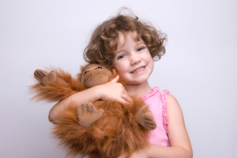 A young child with curly hair, wearing a pink sleeveless top, smiles while hugging a large, fluffy brown stuffed orangutan against a plain light background.