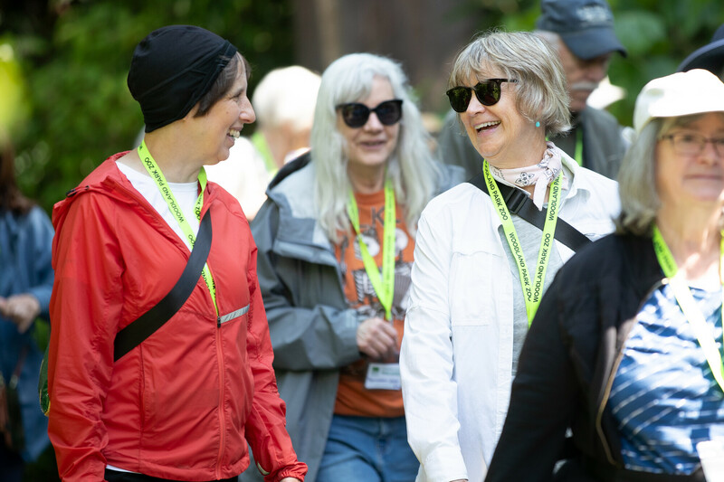 A group of smiling adults wearing bright green lanyards walks outdoors. Two women in front, one in a red jacket and one in a white jacket with sunglasses, appear to be enjoying a conversation.