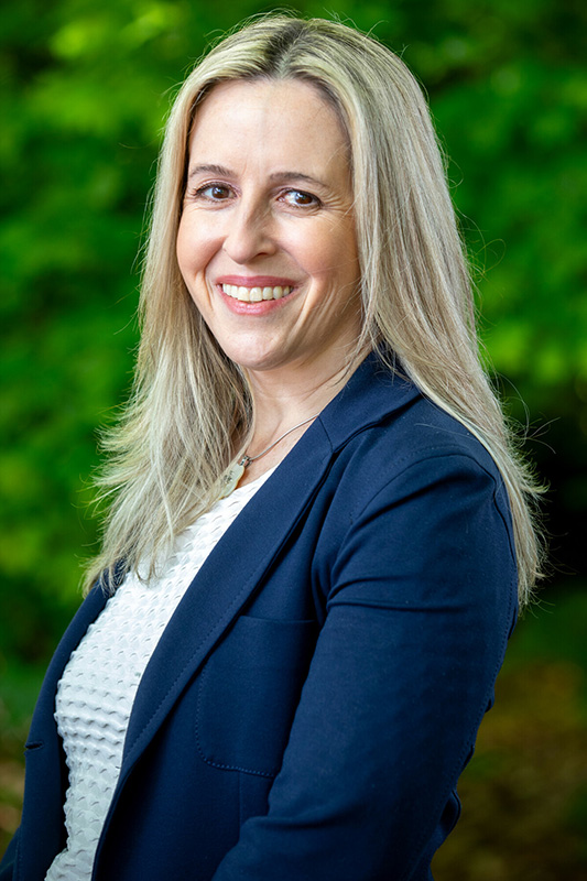 A woman with long blonde hair, wearing a navy blue blazer and white top, smiles at the camera with a blurred green outdoor background.