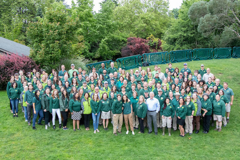 A large group of people, mostly wearing green shirts or jackets, stand together on a grassy lawn surrounded by trees and greenery, posing for a group photo outdoors.