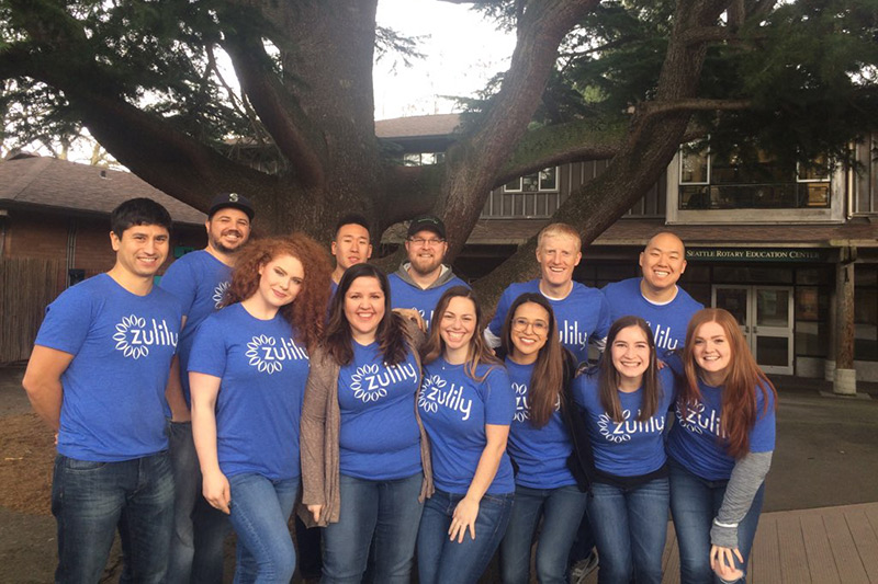 A group of thirteen people wearing matching blue Zulily t-shirts stand and kneel together, smiling, in front of a large tree and a building with a sign that reads “Seattle Hebrew Education Center.”.