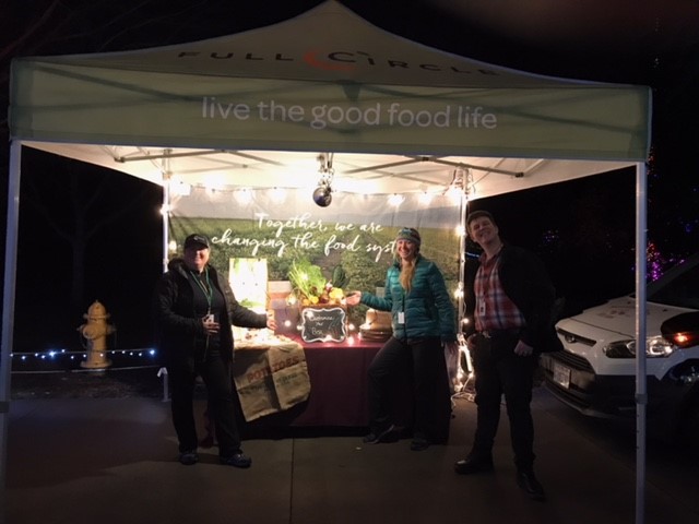 Three people stand smiling under a canopy tent with live the good food life written on it. The table beneath the tent displays produce and a chalkboard sign, with string lights creating a warm, inviting atmosphere.