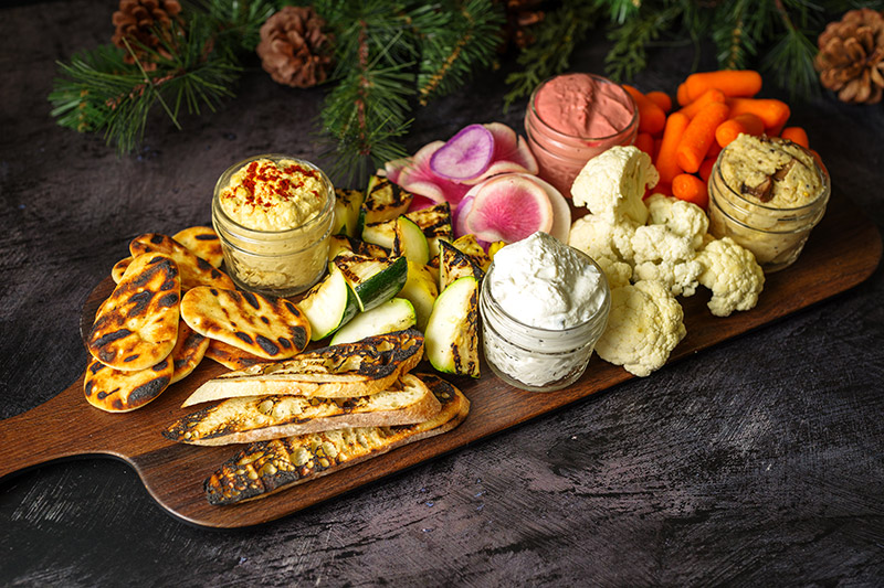 A wooden board with grilled bread, pita, zucchini, hummus, creamy dips, radishes, baby carrots, and cauliflower, set against a dark background with pine branches and pinecones.