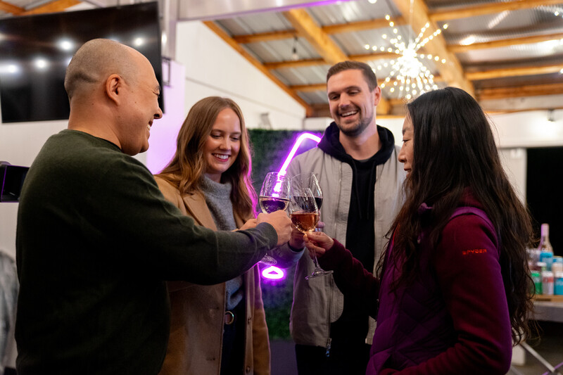 Four people stand in a semi-circle indoors, smiling and clinking glasses of wine in a celebratory toast. Warm lighting and a neon sign add to the festive atmosphere.