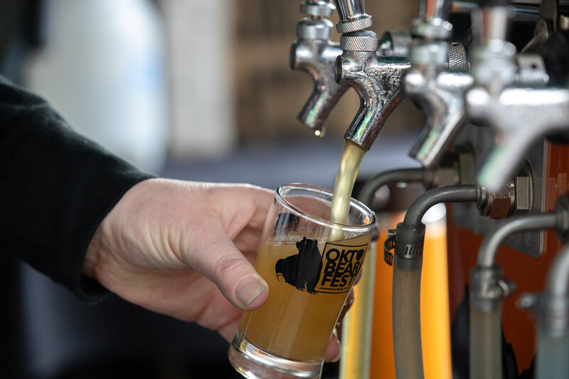 A hand holds a glass labeled Oktobearfest under a beer tap, filling it with light-colored beer at a festival or bar setting.