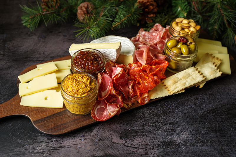 A wooden platter with assorted cheeses, cured meats, crackers, jars of mustard, fig jam, olives, and almonds, set on a dark surface with pine branches in the background.