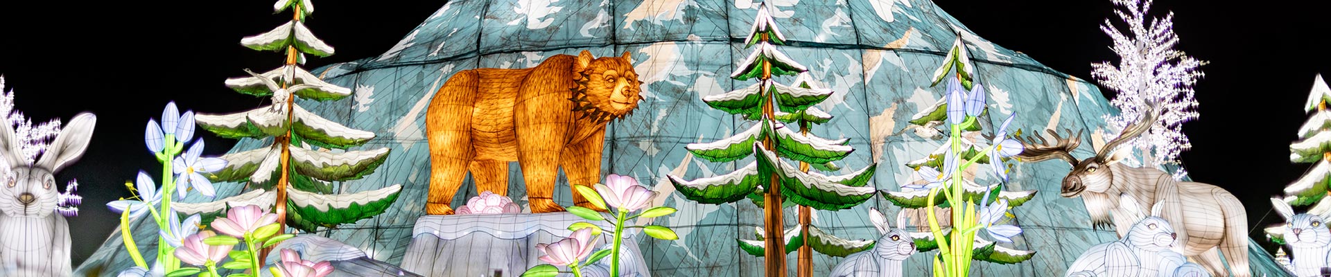 A nighttime scene with illuminated animal lanterns, including a bear, moose, rabbits, and an owl, surrounded by glowing trees and flowers in front of a mountain backdrop.