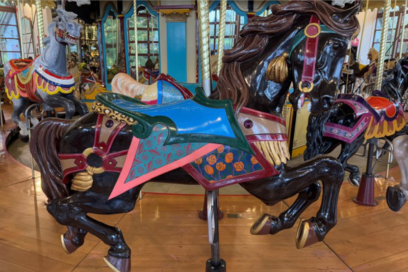 A brightly painted black carousel horse with a colorful saddle, harness, and shield stands on a wooden platform, surrounded by other carousel horses and mirrors in the background.