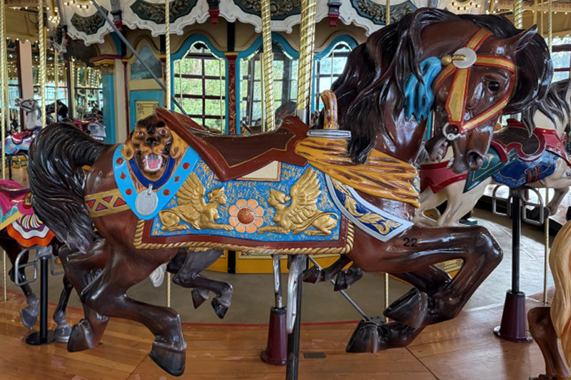 A colorful carousel horse with a painted blue, gold, and red saddle and a lion’s head detail stands on a wooden platform among other carousel animals.