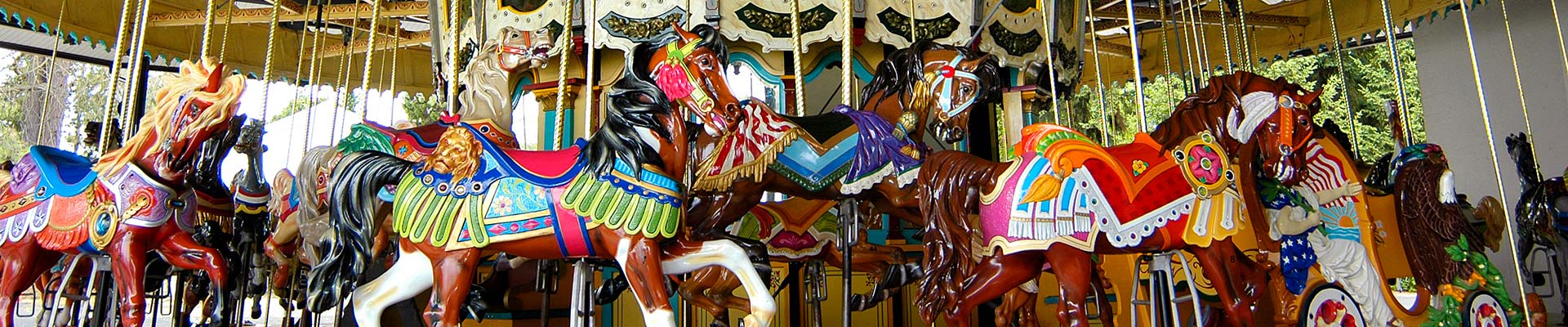 A close-up view of a colorful carousel with intricately painted horses and decorative details, set outdoors with trees visible in the background.