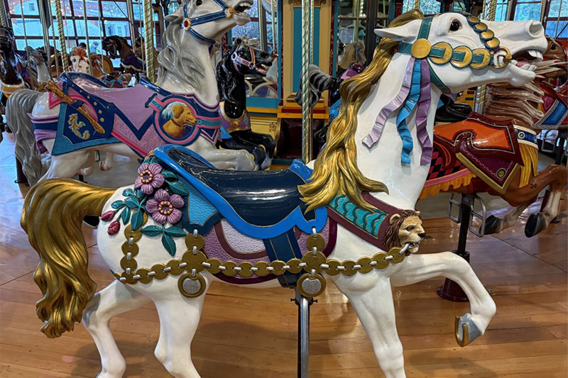 A brightly painted carousel horse with blue and gold saddle, pink flowers, and a golden mane stands indoors among other decorated carousel horses.